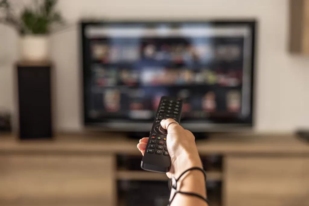 A Woman Is Holding A Remote Control Of A Smart TV In His Hand. In The Background You Can See The Television Screen With Streaming Entertainment Apps For Video On Demand.