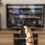 A Woman Is Holding A Remote Control Of A Smart TV In His Hand. In The Background You Can See The Television Screen With Streaming Entertainment Apps For Video On Demand.