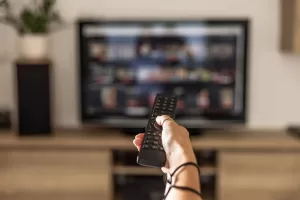 A Woman Is Holding A Remote Control Of A Smart TV In His Hand. In The Background You Can See The Television Screen With Streaming Entertainment Apps For Video On Demand.